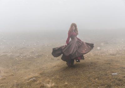 Une femme rousse en robe longue brune et rouge danse dans un paysage brumeux. automnal
