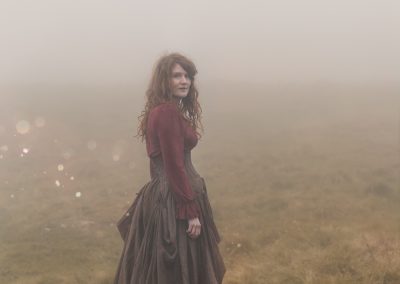 Une femme rousse en robe longue brune et rouge style écossaisese tient dans un paysage brumeux.