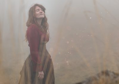 Une femme en robe longue brune et rouge se tient dans un paysage brumeux.