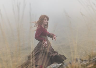Une femme rousse en robe longue brune et rouge danse dans un paysage brumeux. automnal